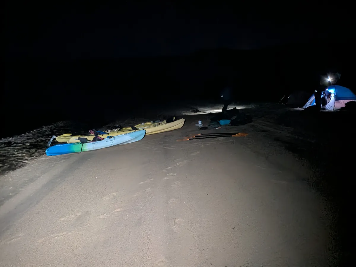 Kayaks and paddles on the beach at night, tents lit by headlamps